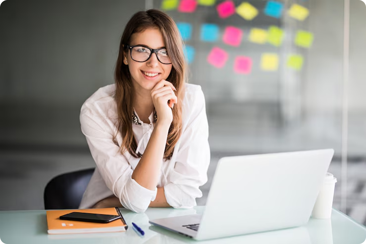 Mulher jovem de óculos sorrindo em um ambiente de escritório moderno com laptop e post-its ao fundo