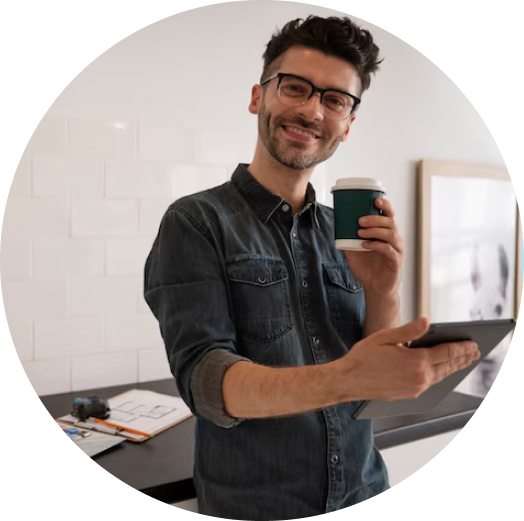 Homem jovem sorridente de óculos, vestindo camisa jeans, segurando um tablet e um copo de café