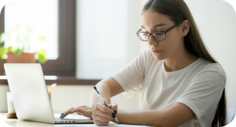 Estudante de óculos concentrada escrevendo em seu caderno enquanto utiliza um laptop