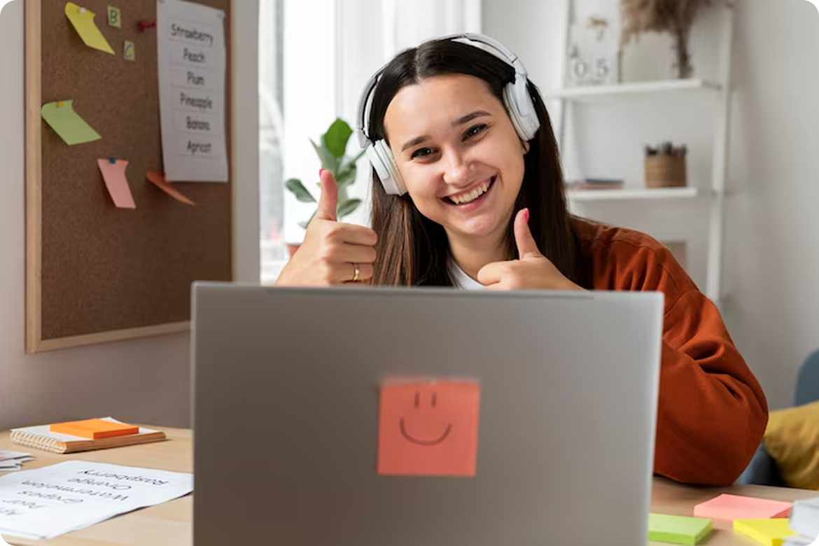 Estudante sorridente com fones de ouvido fazendo sinal de positivo em frente ao laptop