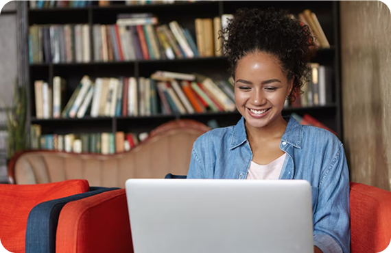 Estudante negra sorridente com cabelo cacheado preso, utilizando um laptop em uma biblioteca com estantes repletas de livros ao fundo